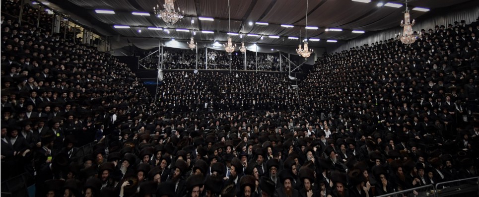 A large Viznitz gathering beneath chandeliers, with people filling the hall and galleries